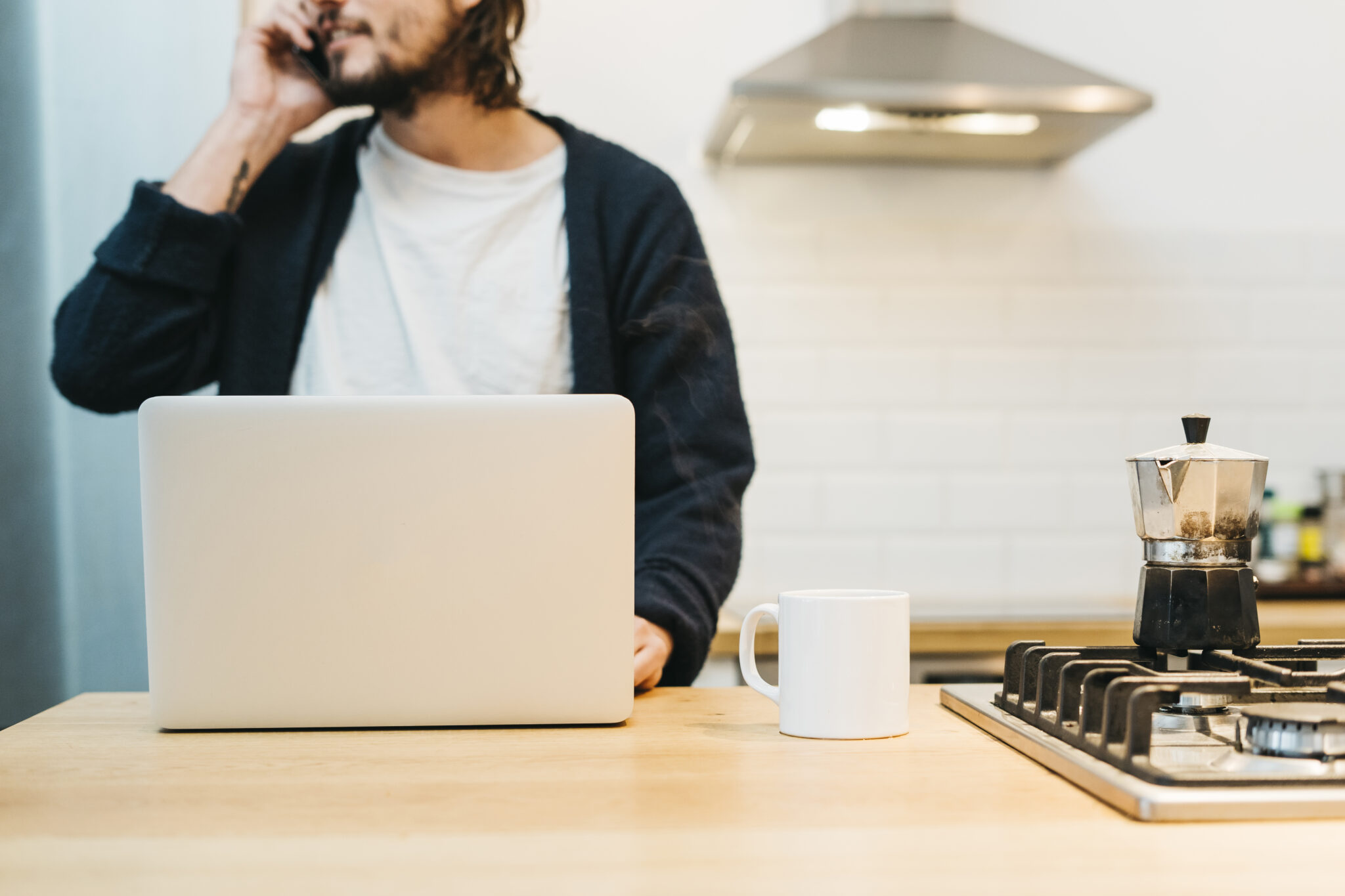 close up man talking mobile phone with laptop white mug kitchen counter scaled